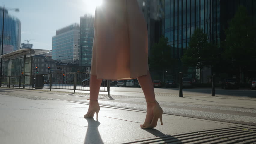 Side view following young woman steps walking city street on sunny summer day. Female wearing casual dress and high heels goes on urban sidewalk. Low angle slim legs backlit warm sunlight, slow motion