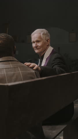 Vertical shot of elderly priest sitting on bench in church, holding hand on shoulder of parishioner and giving him advice