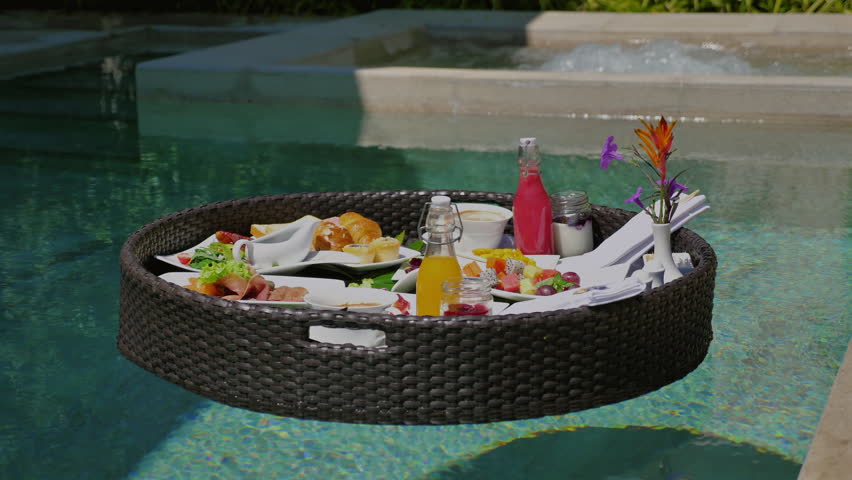 Floating breakfast tray in luxury pool with tropical flowers, fresh fruits, croissants, coffee and juice. Elegant tropical resort amenity and dining experience.