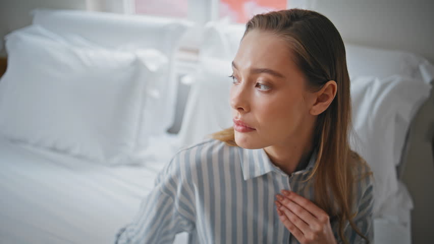 Pensive woman resting on bed feeling sadness closeup. Portrait of beautiful girl in striped pajama deep in thought sitting cozy white bedroom alone. Depressed lady enjoy comfortable home environment.