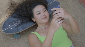 A woman with long hair lies on her back on a skateboard, looking at her phone. She is wearing a green tank top and jewelry, appearing content and engaged with her device taking selfies - Powered by Shutterstock - Get 15% off with code: PIKWIZARD15