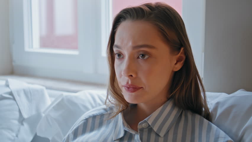 Sad woman sitting morning bedroom radiating solitude closeup. Pensive girl lying white pillows bed reflecting deep thoughtfulness. Portrait of melancholic attractive lady unmotivated to day start.