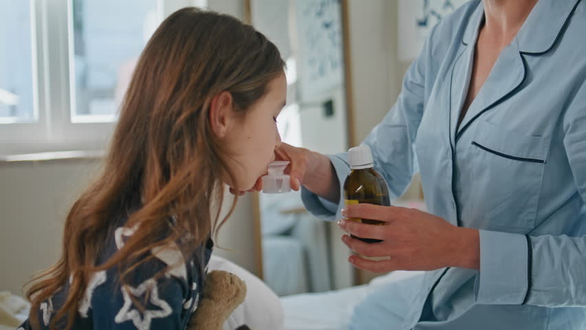 Woman giving mixture to sick daughter at white bedroom closeup. Unrecognizable mom in pyjama sitting bed spending time together with ill child. Small kid taking medicine at bedding. Family concept