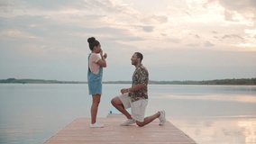 Side vie of African American couple on pierce of large lake. Man standing on knee and proposing to beautiful girl. After saying yes he hugs her and starts swinging around with joy and happiness. - Powered by Shutterstock - Get 15% off with code: PIKWIZARD15