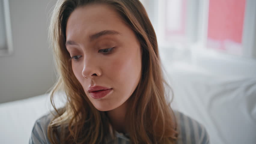 Pensive lady sitting bed early morning showing concerned expression closeup. Serious pretty girl feeling loneliness in cozy white bedroom. Sad woman thinking on problems at modern apartment alone.