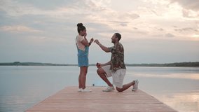 Stable view of African American couple standing at end of pierce. male kneeling in front of woman and proposing to her. Woman with shock taking ring and coming to hug her beloved man. Proposal concept - Powered by Shutterstock - Get 15% off with code: PIKWIZARD15