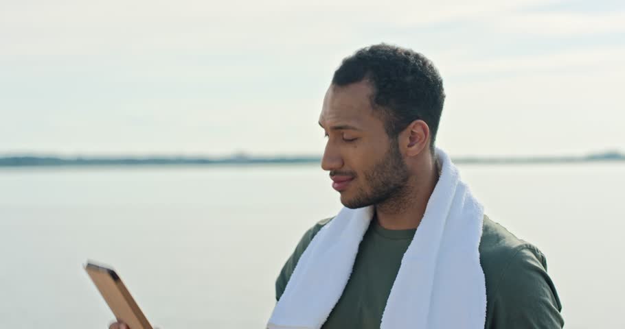 African American male holding his tablet device with one hand. Talking with relatives or friends next to large lake. White towel around his neck. Waving his hand to greet with friends.