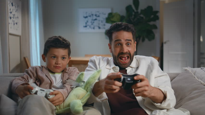 Excited gamers family playing video game at dark home closeup. Cheerful father small son holding controllers enjoying videogame at living room. Emotional man boy using joysticks together at house