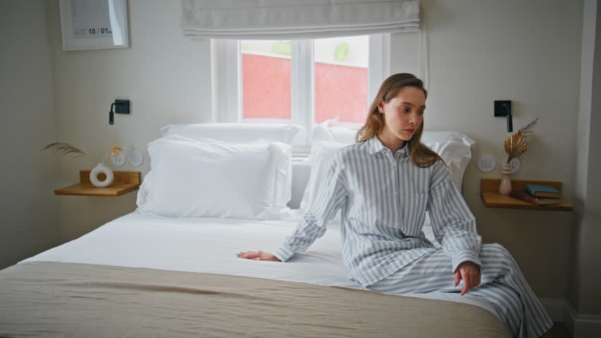 Sad girl sitting bed modern hotel room touching soft white blanket. Upset woman relaxing at cozy bedroom in thoughtful pose. Morning light illuminating pensive expression lady in striped pajamas.