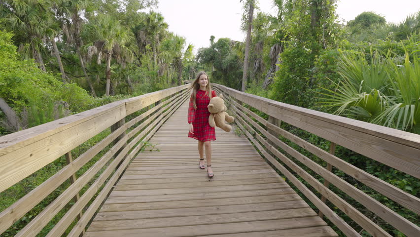 Teenage girl walking alone with her teddy bear on wooden boardwalk trail through wild tropical jungles. Child exploring Florida nature in dense green rainforest.