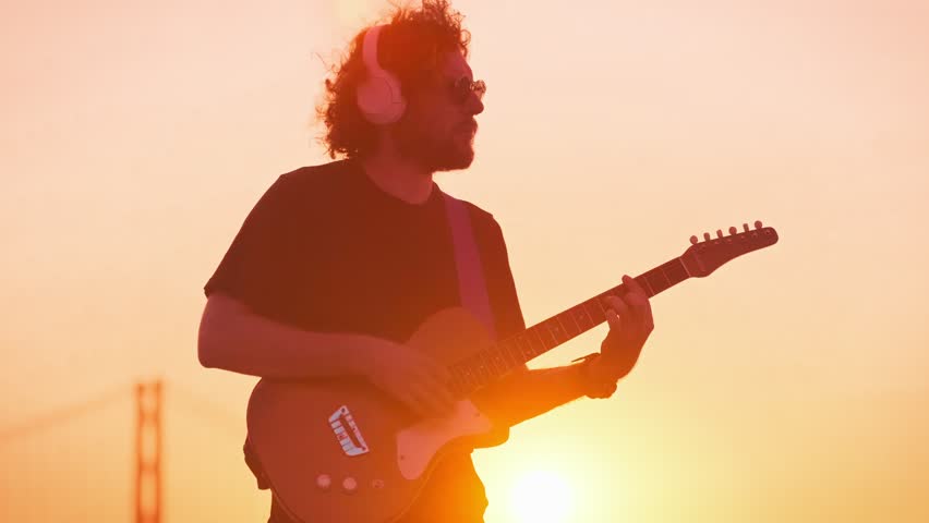 Hipster street musician in black playing electric guitar in street outdoors on sunset silhouette