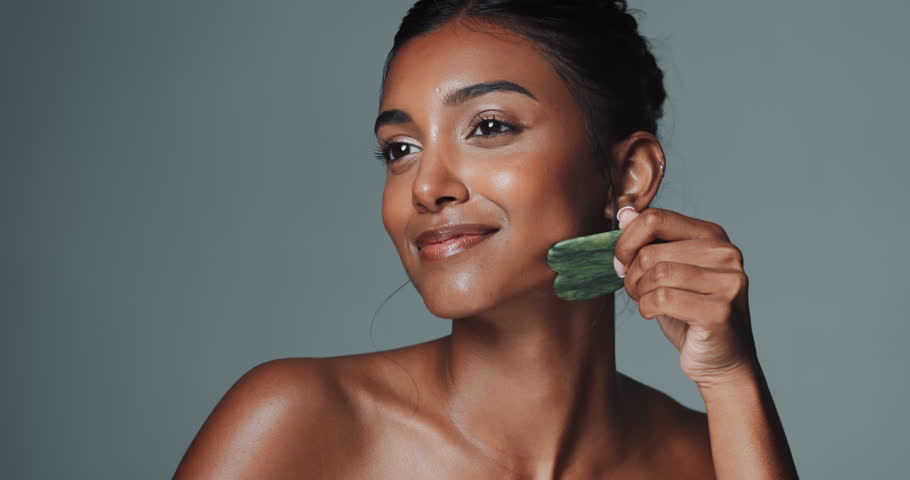 Woman, studio and happy with face guasha, cosmetic and massage for facial structure on background. Technique, skincare and dermatology for beauty tool, glow and natural jade stone for Chinese therapy