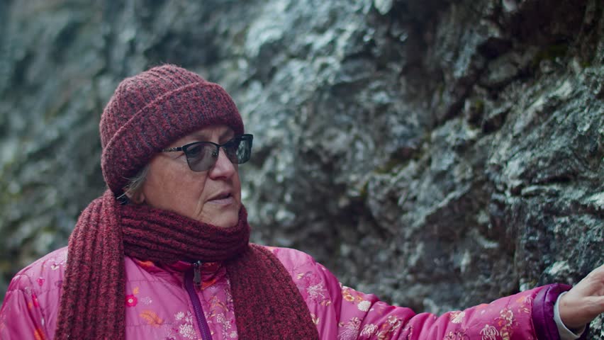 An elderly woman in glasses and a puffer jacket walks in the gorge, traveling and inspecting autumn rocks