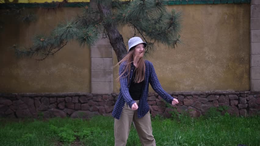 young woman in a blue plaid shirt and beige pants dances hip-hop energetically in front of a yellow wall with stone accents. She wears a white bucket hat and black shoes