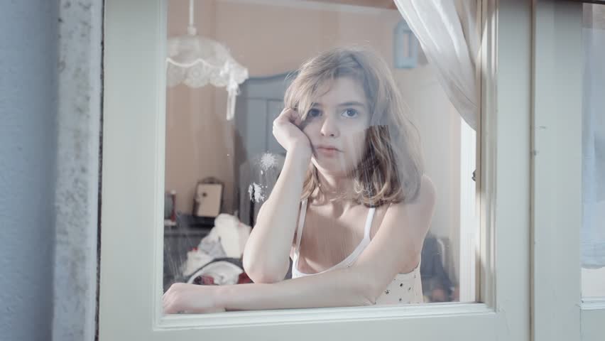 A little girl, looking sad and bored, gazes out the window. Seen from the front, her expression is visible through the glass.
