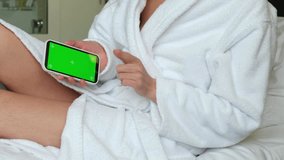 A woman in a bathrobe is sitting on the bed. Women's hands will scroll the phone with a green screen. 4k video. High quality 4k footage