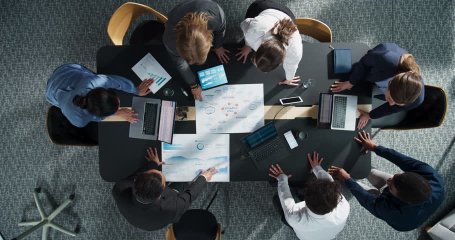 Top Down View Of Diverse Team Of Business Professionals And Managers With Laptops Entering The Conference Room And Sitting Behind Table To Discuss Financial Data And Sales Reports In Corporate Office.