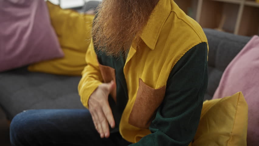 A redhead bearded man in a yellow shirt holding his chest feels discomfort on a sofa indoors.