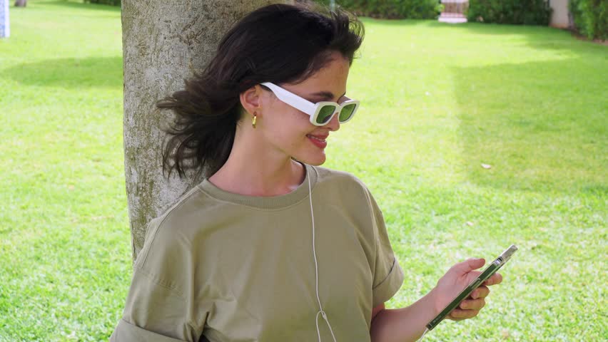 A Young Woman With Loose Brown Hair, Wearing A Green T-Shirt, White Sunglasses, White Earphones, And Gold Earrings, Putting On An Earphone And Laughing While Checking Her Phone. Leaning Against A Tree