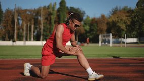 Warm-up for the knee joints of a male athlete as he trains outdoors on a sunny autumn day on the stadium running track. A young athlete with dreadlocks and wearing sunglasses does stretching at the - Powered by Shutterstock - Get 15% off with code: PIKWIZARD15
