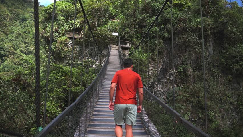 hispanic tourist, seen from behind, crosses a wooden hanging bridge in front of the majestic Pailon del Diablo waterfall in Baños de Agua Santa, Ecuador
