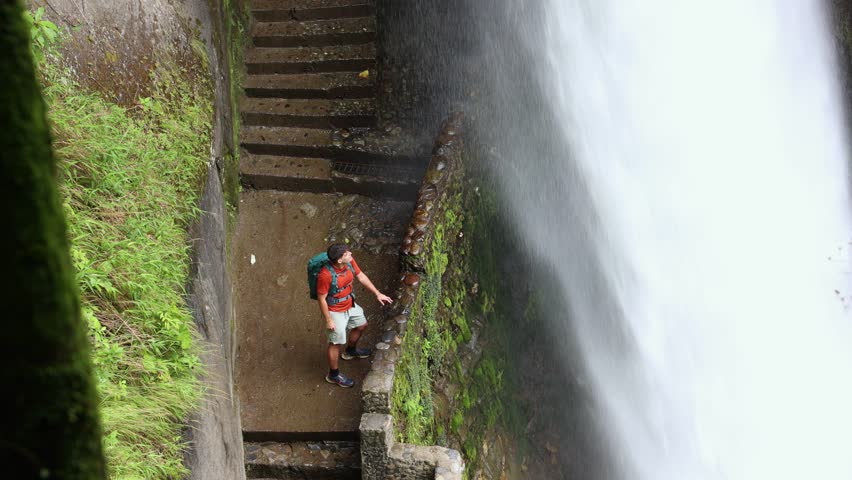 Latin traveler with a backpack stands close to Pailon del Diablo waterfall in Baños de Agua Santa, Tungurahua, Ecuador, observing the powerful force of the water up close