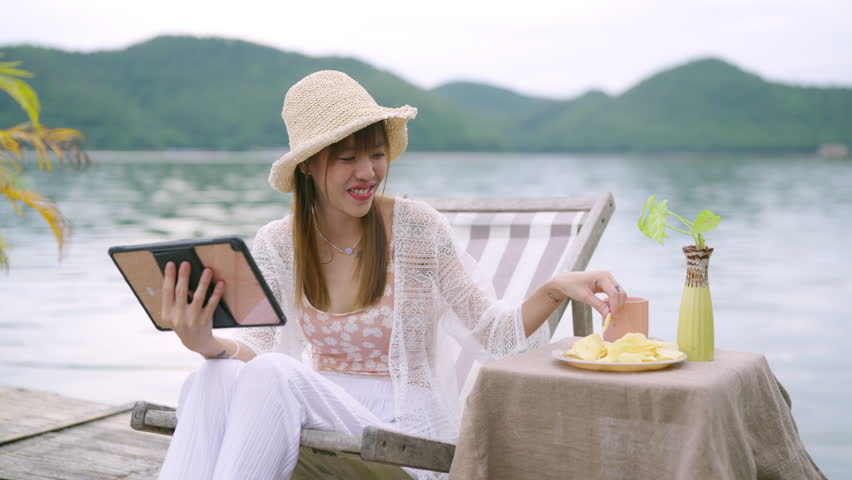 Asian woman sitting on lake house balcony and using digital tablet remote working online business or watching movie. Attractive girl enjoy outdoor lifestyle travel nature on summer holiday vacation.