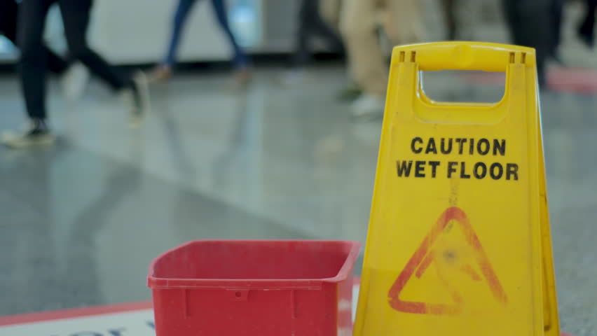 A wet floor and bucket warning sign with many train passengers' feet in the background walking and passing by on the corridor platform of a commuterline electric train station at rush hour