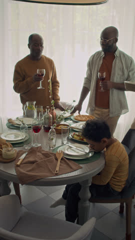 Full vertical slowmo of African American family sitting down to festive dinner at home, mother and daughter bringing trays with hot food, while father and granddad are tasting homemade wine by window