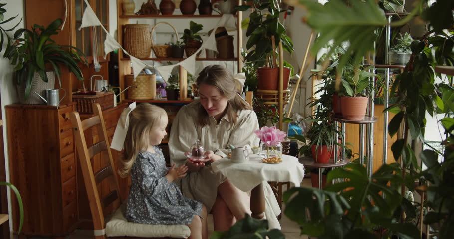 Young loving mother drinking tea with her adorable little daughter in grey dress among decorative plants in the home garden. Family joint leisure and care.