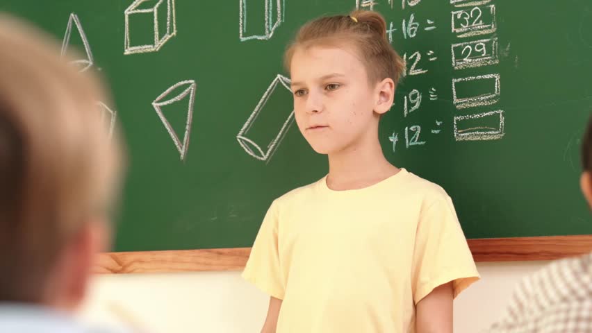 Caucasian boy talking and present idea while standing in front of blackboard with Math theory written. Diverse cute student sitting while looking at new student introduce himself. Education. Pedagogy.