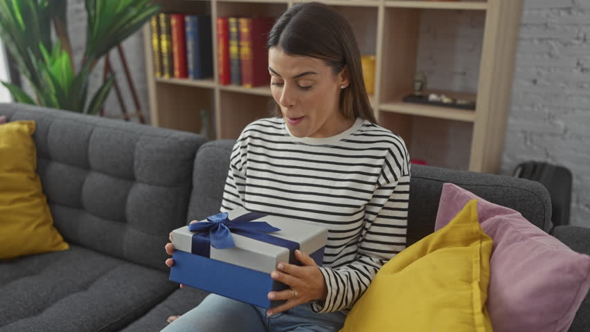 Surprised young hispanic woman opening a blue ribboned gift box in a cozy living room