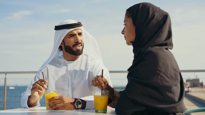 Cinematic image of a young couple from the united arab emirates spending time outdoor. Man and woman with traditional emirati kandura and abaya sitting in a bar and drinking an orange juice.