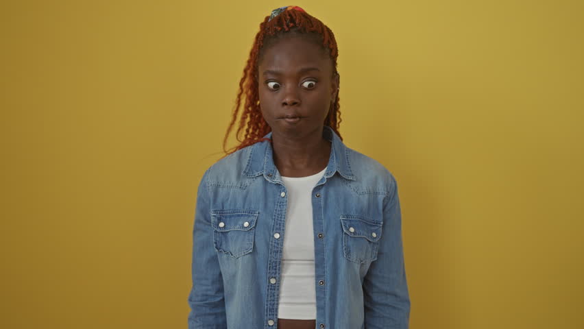 Fun, comical african american woman wearing denim shirt, making a fish face expression; standing over an isolated yellow background showcasing her crazy, funny nick in a silly gesture with lips
