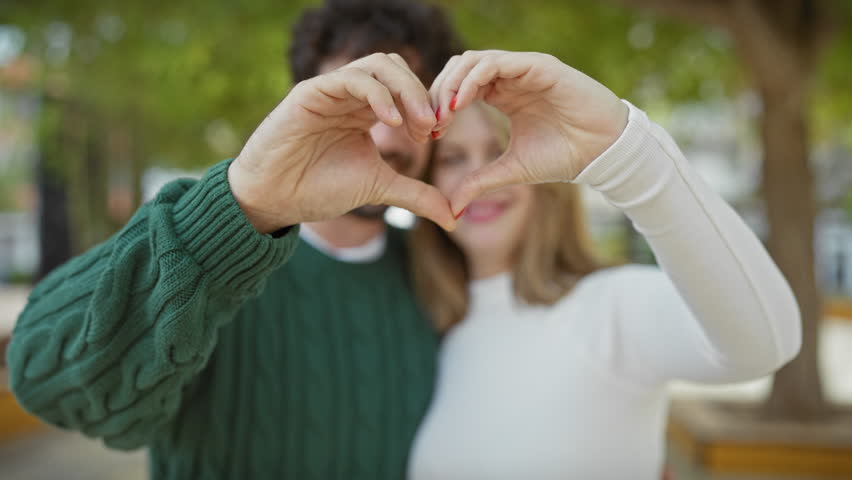 A smiling couple forms a heart shape with their hands in a lush park, symbolizing love and togetherness against a nature-filled backdrop.