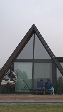 People sit on bench and chair near modern triangle-shaped country house with panoramic windows. Family relaxes in yard on cloudy day Vertical Shot.