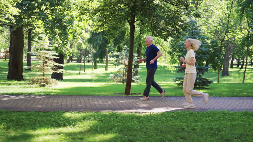 Seniors jogging together on park path surrounded by lush greenery. Concept of active lifestyle, fitness and health in old age. Couple enjoying morning run staying fit and healthy