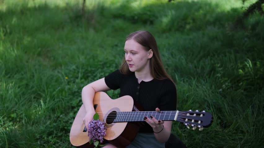 young woman with long hair, dressed in a black top with bouquet of lilac flowers and acoustic guitar. She is outdoors, surrounded by lush green grass and trees, creating a peaceful, natural ambiance