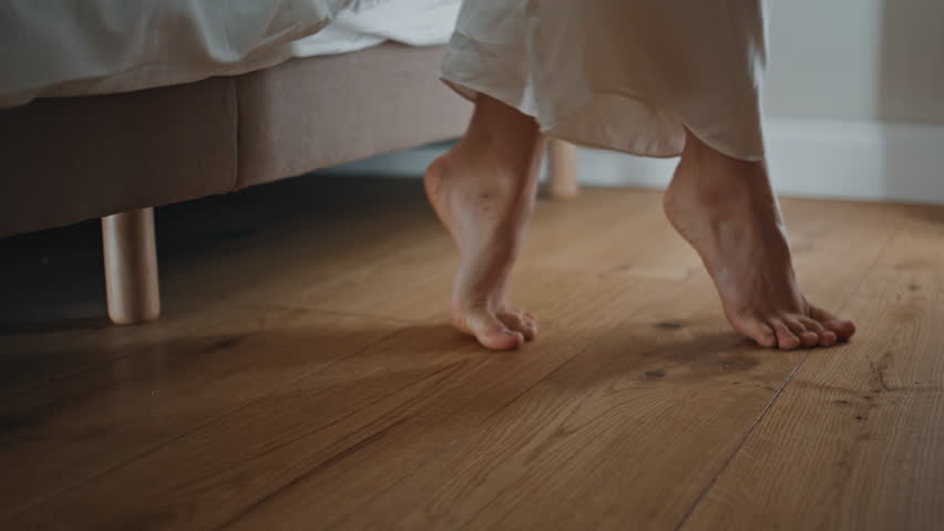 Closeup woman walking toes in morning. Girl waking up get out cozy bed at home. Unrecognizable model going floor in softly lit bedroom. Tranquil person in pajamas stepping barefoot in hotel room.