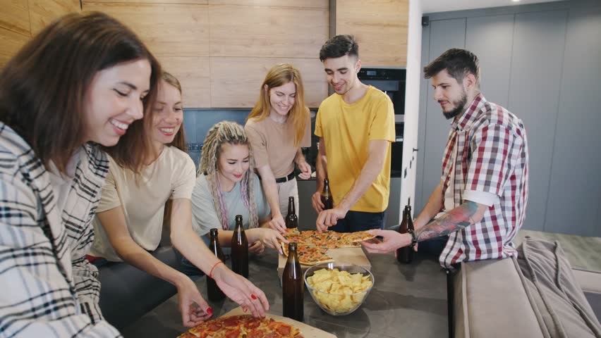 A group of friends at a home party with beer in their hands, take a slice of pizza, eating it and talking