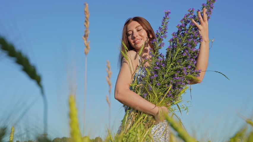 Beautiful girl walking through the lavender field. Close-up portrait of beautiful young lady holding bouquet of salvia sage wild flowers walking in the grass meadow on a summer day.