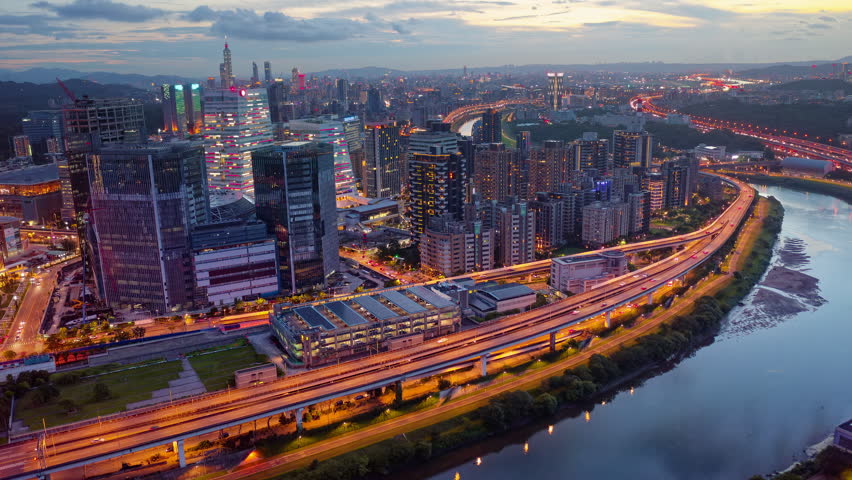 Aerial hyperlapse of Huan Dong Boulevard, an elevated expressway winding along Keelung River in Nangang District, Taipei, with city lights dazzling at dusk and 101 Tower standing out in background