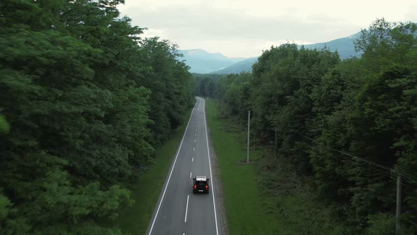 A black SUV drives along the road through a forest belt, mountains are visible in the distance