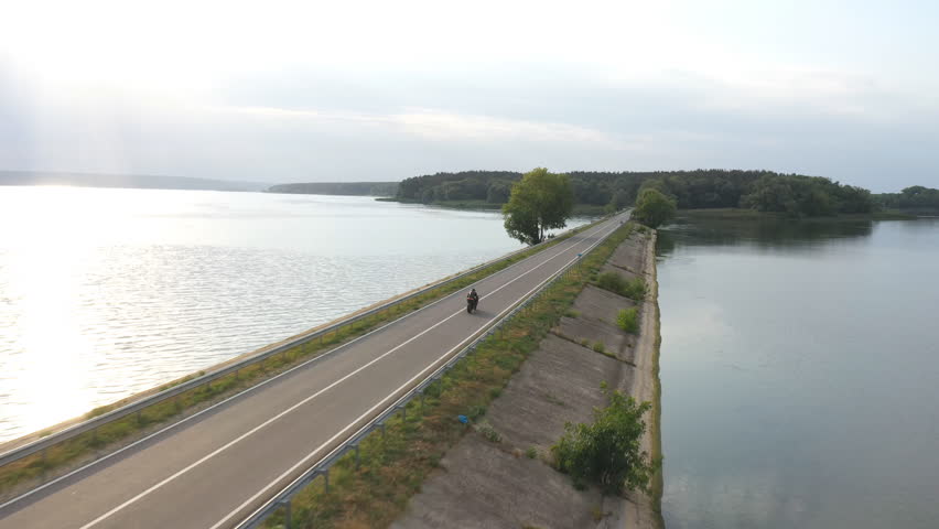 Aerial shot of biker ride on motorbike through road near lake. Motorcyclist racing his motorcycle at dam route on summer day. Man drive bike through bridge of river or pond. Concept of travel.Top view