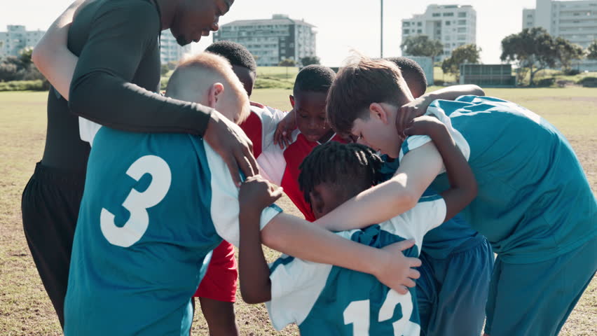 Coach, children and team with huddle for soccer, motivation or game plan with diversity on field. Man, football and group in scrum with support, connection and care with hug for solidarity on pitch
