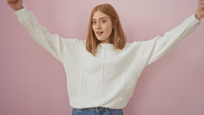 Smiling young redhead woman confidently standing isolated over pink background, wearing sweater, flexing her strong arms muscles, embodying a successful fitness lifestyle concept.