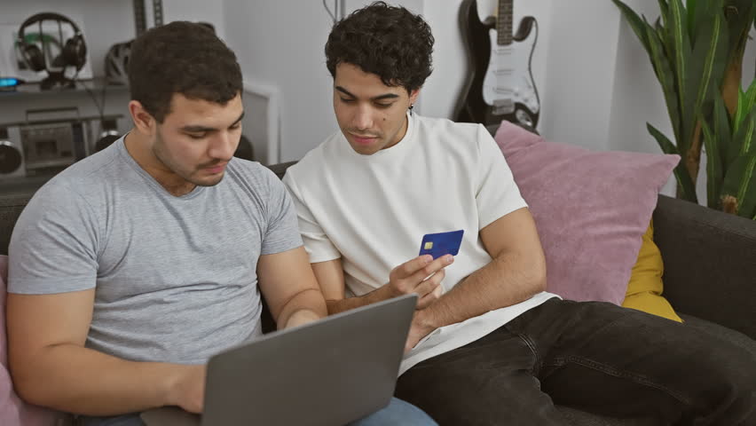 Two men using a laptop and celebrating with a credit card on a sofa at home.