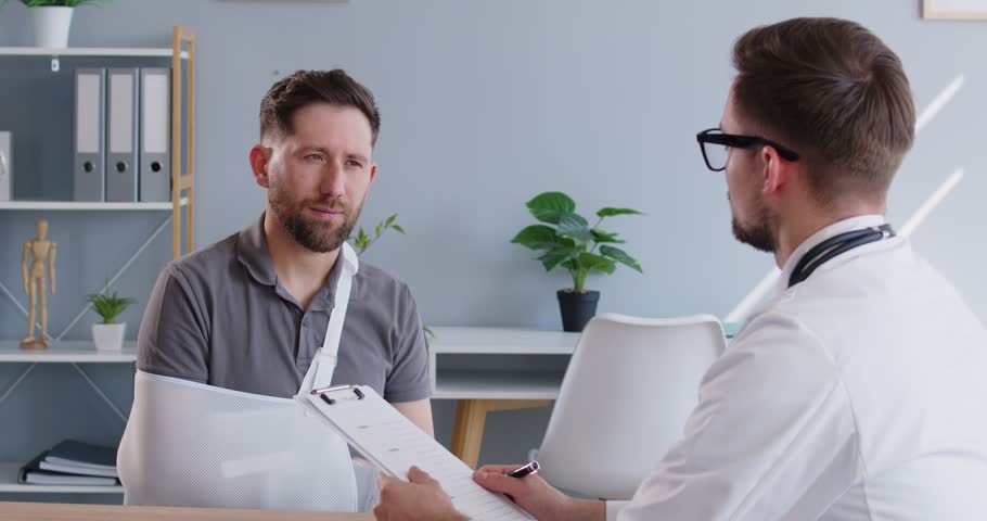 Young male patient with broken arm visiting doctor for consultation about therapy treatment in medical office. Man with injured hand sitting at the desk with traumatologist. 4k video.