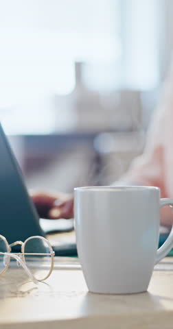 Coffee, glasses and steam with a cup on an office desk while a business person is working in the background. Tea, cup or mug with an employee typing on a laptop in a workplace for communication