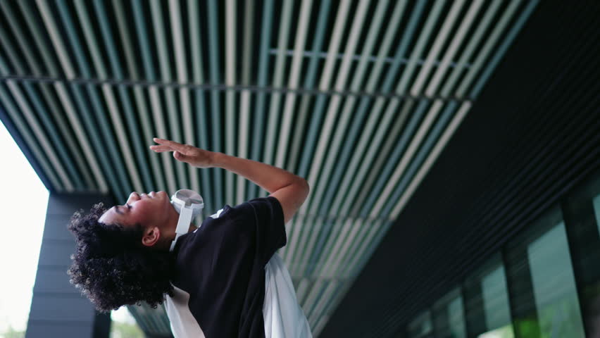 Young smiling dark skinned female with headphones dances outdoors in city environment. Womens joyful expression and casual outfit embody carefree spirit and positive vibes of urban youth culture